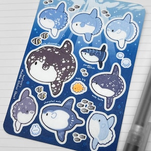 Könnte beinhalten: Ein Stickerbogen mit Cartoon-Illustrationen von Mondfischen in Blau-, Weiß- und Lilatönen. Der Bogen enthält den Text "Mola Mola Sticker Sheet" und "heyitsriin.etsy.com". Ein durchsichtiger Stift liegt auf dem linierten Papier.