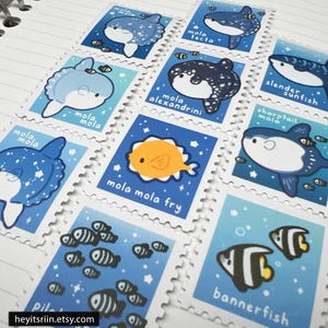 Peut inclure: Une collection de timbres illustr&eacute;s repr&eacute;sentant diverses cr&eacute;atures marines, dont des poissons-lunes et des poissons-drapeaux. Chaque timbre a un fond bleu avec des bordures blanches et les noms des poissons. Les timbres sont dispos&eacute;s sur une surface blanche lign&eacute;e.