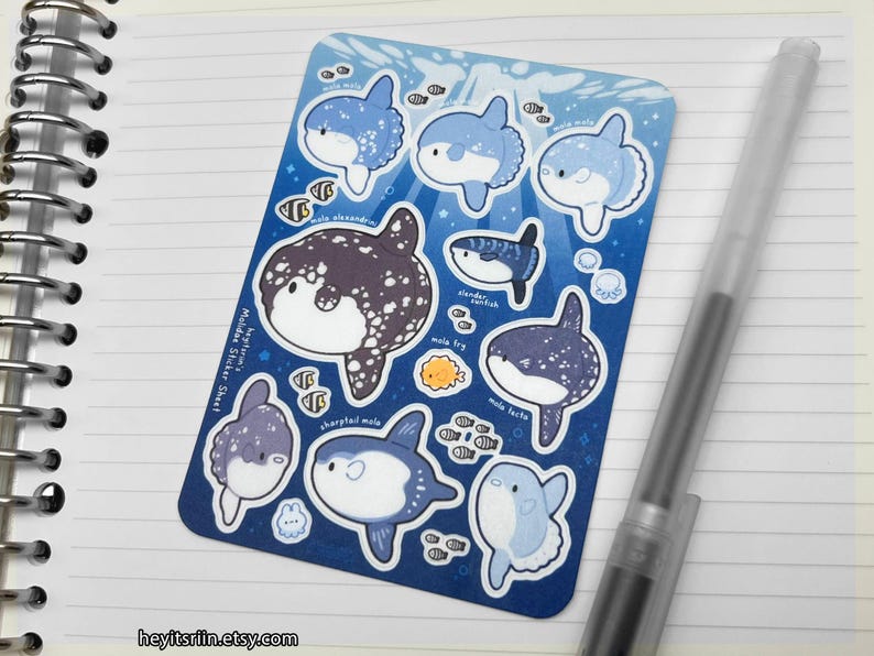 Peut inclure: Une planche d'autocollants avec des illustrations de poissons-lunes de dessins anim&eacute;s en bleu, blanc et violet. La feuille comprend le texte "Mola Mola Sticker Sheet" et "heyitsriin.etsy.com". Un stylo transparent repose sur le papier lign&eacute;.