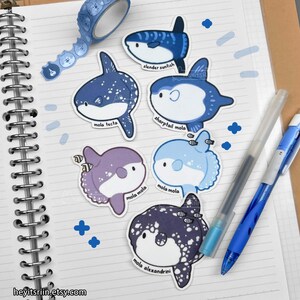 Peut inclure: Plusieurs autocollants de poissons-lune bleus et violets, avec les noms des esp&egrave;ces imprim&eacute;s. Un rouleau de ruban adh&eacute;sif washi bleu &agrave; motifs, deux stylos et des autocollants bleus en forme de croix sont &eacute;galement visibles sur un carnet ouvert.