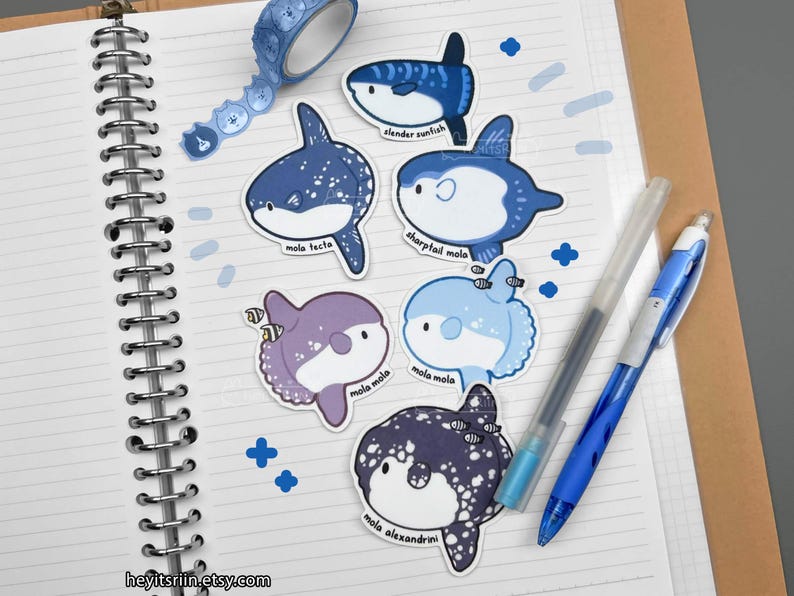 Peut inclure: Plusieurs autocollants de poissons-lune bleus et violets, avec les noms des esp&egrave;ces imprim&eacute;s. Un rouleau de ruban adh&eacute;sif washi bleu &agrave; motifs, deux stylos et des autocollants bleus en forme de croix sont &eacute;galement visibles sur un carnet ouvert.