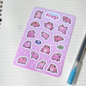 May include: A sheet of pink and white stickers featuring a variety of pink frogs in different poses. The sheet is titled "Frogs" and has a small star design.