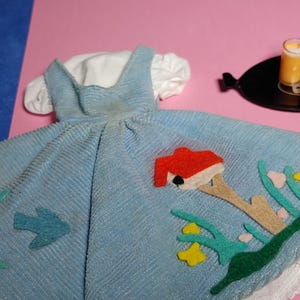 May include: A light blue corduroy dress with a white bodice and a mushroom applique. The dress has a white lace trim and features a green and yellow floral design.