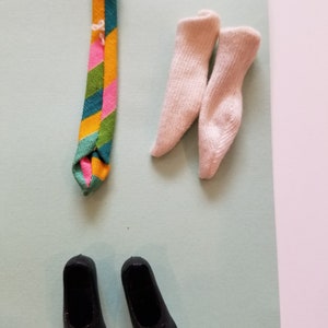 May include: A collection of doll accessories, including a colorful striped tie, two pairs of light pink socks, and two black shoes. The tie features stripes of green, yellow, pink, and blue. The socks are a light pink color.