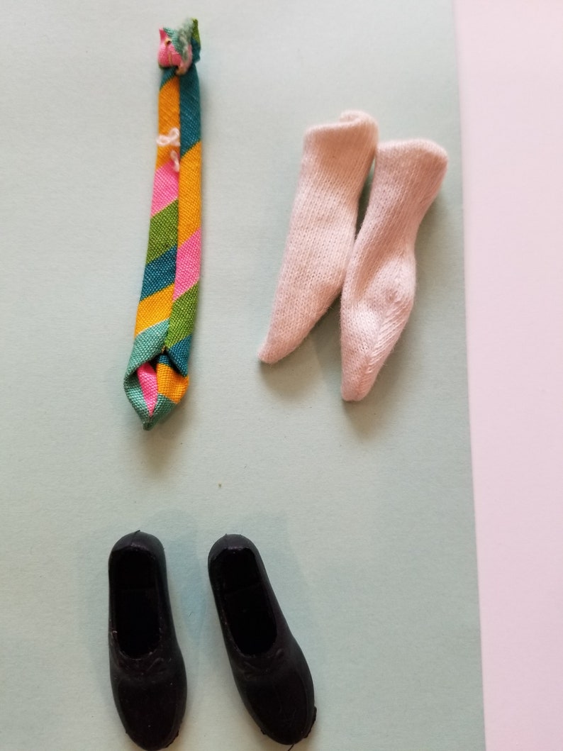May include: A collection of doll accessories, including a colorful striped tie, two pairs of light pink socks, and two black shoes. The tie features stripes of green, yellow, pink, and blue. The socks are a light pink color.