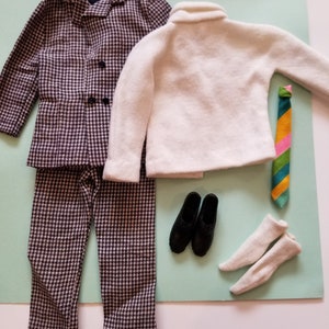 May include: A vintage doll's outfit featuring a houndstooth patterned suit, a white sweater, a striped tie, black shoes, and white socks. The suit includes a jacket and pants. The ensemble is arranged on a light green surface.