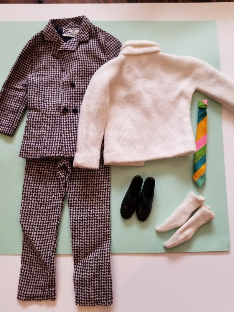May include: A vintage doll's outfit featuring a houndstooth patterned suit, a white sweater, a striped tie, black shoes, and white socks. The suit includes a jacket and pants. The ensemble is arranged on a light green surface.