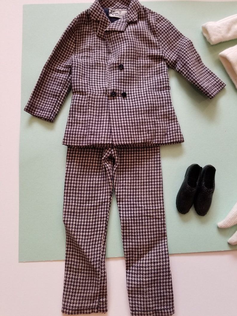 May include: A miniature houndstooth suit set, including a jacket with two black buttons and matching pants. The suit is paired with black shoes. The suit is in a black and white pattern.