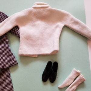 May include: A miniature doll outfit featuring a white sweater, black shoes, white socks, and a houndstooth patterned coat. The items are arranged on a light green surface, suggesting a fashion theme.