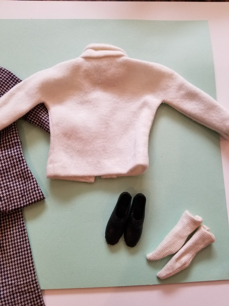 May include: A miniature doll outfit featuring a white sweater, black shoes, white socks, and a houndstooth patterned coat. The items are arranged on a light green surface, suggesting a fashion theme.