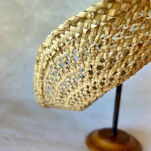 May include: A woven straw hat with a wide brim, displayed on a dark metal stand with a wooden base. The hat's weave creates an open, airy pattern, and the natural straw colour is prominent.
