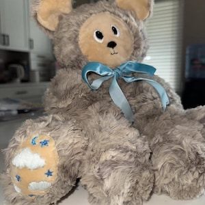 May include: A plush teddy bear with a mouse-like appearance. The bear is primarily gray with tan accents on the ears and face. It has a blue ribbon tied around its neck and a paw with a moon and star design.