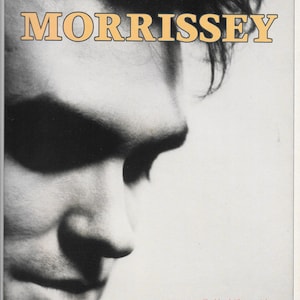 Op de afbeelding: Zwart-wit vintage advertentie met Morrissey. De afbeelding toont een close-up van het gezicht van een man met de naam "MORRISSEY" in een bruinachtige lettertype bovenaan. Hieronder staat de tekst "Viva Hate" en "The Album, Compact Disc, Cassette."