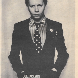 May include: Vintage advertisement for Joe Jackson's debut album, "Look Sharp!" The black and white image features a man in a suit, striped shirt, and polka dot tie. Text includes "Joe Jackson Looks Sharp" and album details.