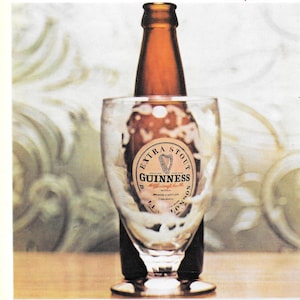 May include: A brown Guinness Extra Stout bottle is partially inserted into a clear glass. The glass is filled with a dark liquid and foam. The Guinness label is visible, featuring a harp symbol.