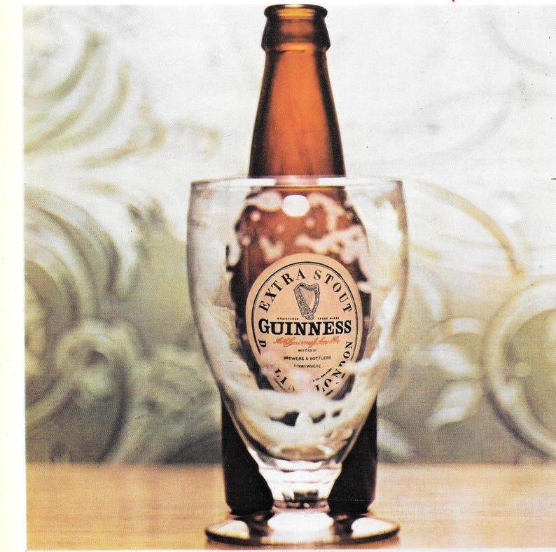 May include: A brown Guinness Extra Stout bottle is partially inserted into a clear glass. The glass is filled with a dark liquid and foam. The Guinness label is visible, featuring a harp symbol.
