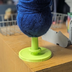 Op de afbeelding: Een blauwe bol garen wordt op een groene garenkom met een witte haak gehouden.