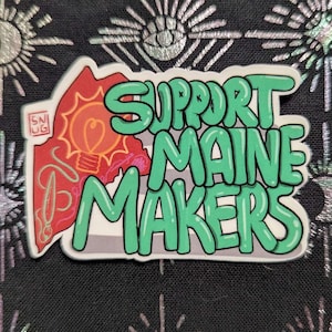 Könnte beinhalten: Ein Aufkleber mit dem Text "SUPPORT MAINE MAKERS" in grünen Buchstaben. Der Aufkleber zeigt einen roten Umriss des Bundesstaates Maine mit einem Glühbirnen-Design. Der Hintergrund ist schwarz mit einem silbernen Muster.