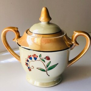 以下が含まれることがあります： 蓋と2つの取っ手が付いたヴィンテージの砂糖入れ。ボウルはクリーム色で、花柄と金色のアクセントが付いています。蓋と取っ手は金色です。花柄は赤と青の花と緑の葉が特徴です。