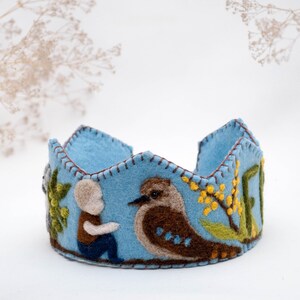 May include: A blue felt crown with a stitched design featuring a bird, a person, and flowers. The crown is decorated with brown, green, and yellow felt.