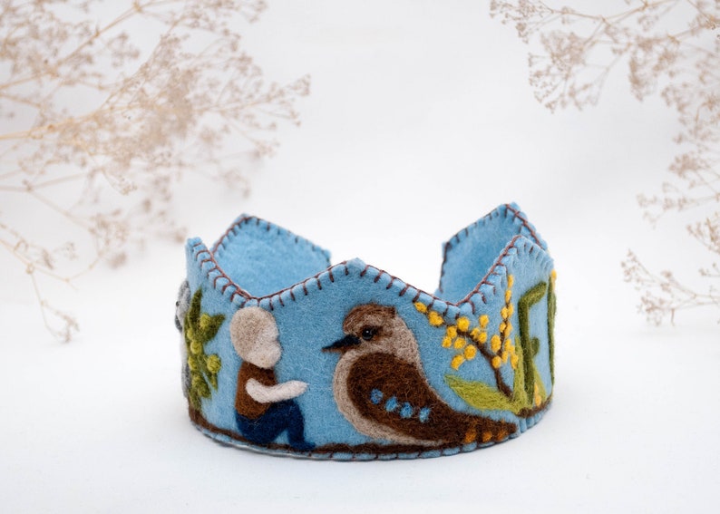 May include: A blue felt crown with a stitched design featuring a bird, a person, and flowers. The crown is decorated with brown, green, and yellow felt.