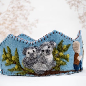 May include: A blue felt crown with a brown felt trim. The crown features two grey felt koalas holding onto a green felt branch with yellow felt leaves. A small brown felt figure is on the right side of the crown.
