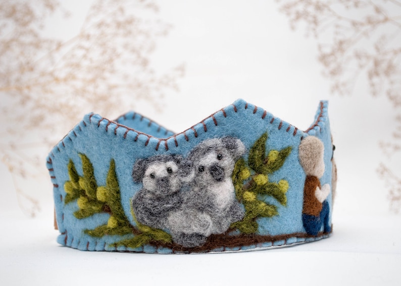 May include: A blue felt crown with a brown felt trim. The crown features two grey felt koalas holding onto a green felt branch with yellow felt leaves. A small brown felt figure is on the right side of the crown.