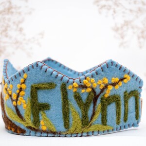 May include: A blue felt crown with the name "Flynn" embroidered in green and yellow felt. The crown is decorated with yellow felt flowers.