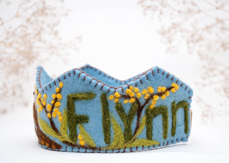 May include: A blue felt crown with the name "Flynn" embroidered in green and yellow felt. The crown is decorated with yellow felt flowers.
