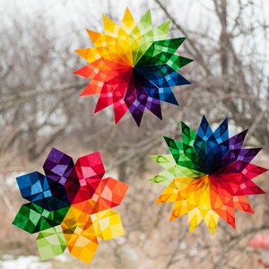 May include: Three rainbow-colored paper stars, each with a different design, are displayed on a window. The stars are made of folded paper and have a geometric pattern.