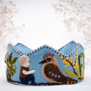 May include: A blue felt crown with a stitched design featuring a bird, a person, and flowers. The crown is decorated with brown, green, and yellow felt.
