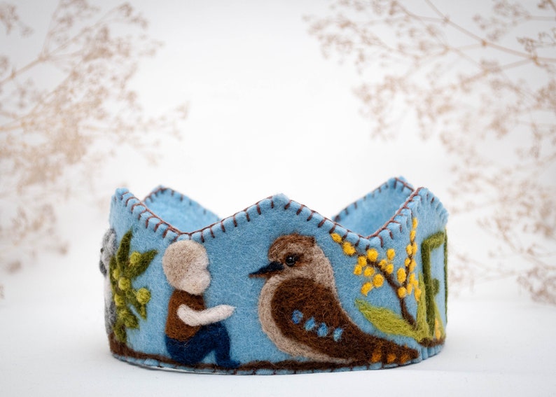 May include: A blue felt crown with a stitched design featuring a bird, a person, and flowers. The crown is decorated with brown, green, and yellow felt.
