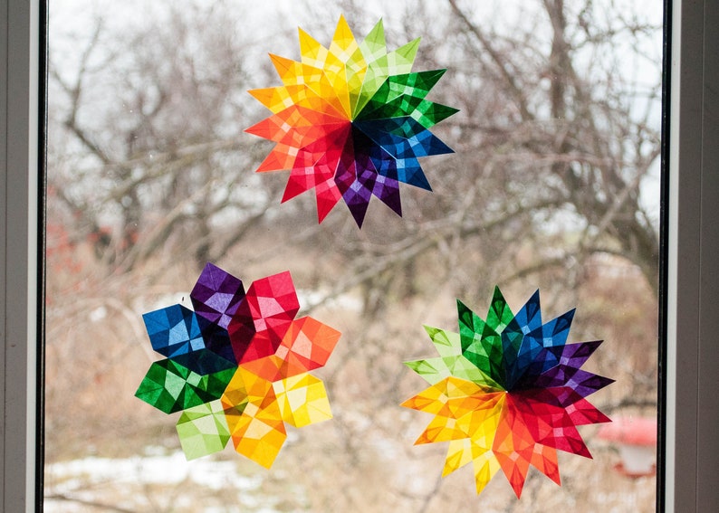 May include: Three colorful paper stars, each with a rainbow pattern, are displayed on a window. The stars are made of tissue paper and are folded into a three-dimensional shape.