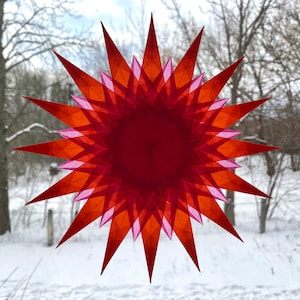 May include: A red and pink paper star with a 3D effect, hanging in a window. The star is made of multiple layers of paper, creating a layered effect.