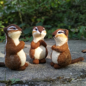 May include: Three handmade felt chipmunk figurines. Each chipmunk has brown fur, a white belly, and a striped back. They are posed with their arms crossed, and tails extended. The chipmunks are standing on a gray surface with a green background.