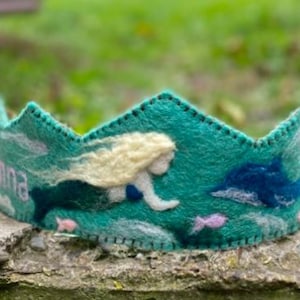 May include: A teal felt crown with a mermaid and dolphin design. The crown features a mermaid with blonde hair, a blue dolphin, and the name "Anna" in pink lettering. The crown is decorated with pink and white accents.