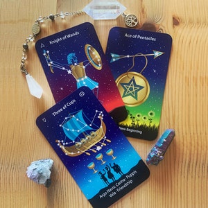 Può includere: Tre carte dei tarocchi con un tema celeste. Le carte sono "Knight of Wands", "Three of Cups" e "Ace of Pentacles". Le carte sono disposte su una superficie di legno con un pendolo di cristallo e un cristallo.