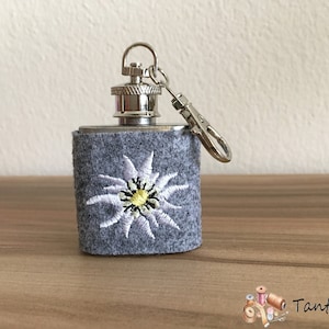 Könnte beinhalten: Eine kleine silberne Trinkflasche mit einem grauen Filzbezug, auf dem eine gestickte weiße Edelweißblume mit gelber Mitte zu sehen ist. Die Flasche hat einen silbernen Verschluss und eine silberne Schlüsselkette.