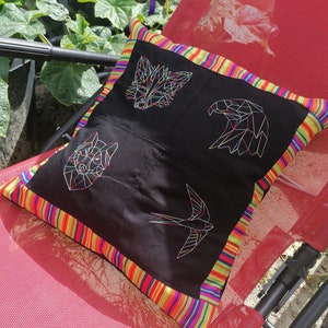 May include: A black pillow with a colorful striped border features geometric embroidered designs of a fox, eagle, bird, and a swallow. The pillow is on a red chair.