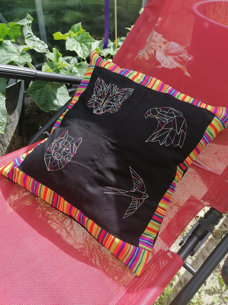 May include: A black pillow with a colorful striped border features geometric embroidered designs of a fox, eagle, bird, and a swallow. The pillow is on a red chair.