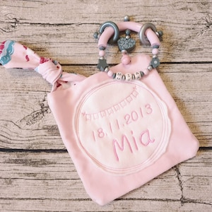 以下が含まれることがあります： ピンクのベビーブランケットに、"18.11.2013 Mia"と刺繍された丸いパッチと、ピンクとグレーのウッドビーズネックレスに「Mia」の文字が書かれています。