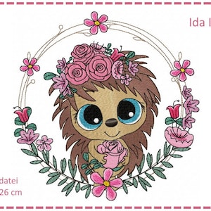 May include: A cute cartoon hedgehog with big blue eyes wearing a pink flower crown and holding a pink rose. The hedgehog is surrounded by a wreath of pink flowers and green leaves. The text "Ida Igel" is written in the upper right corner of the image. The text "Stickdatei 16 x 26 cm" is written in the lower left corner of the image.