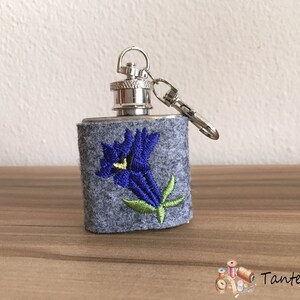 Könnte beinhalten: Eine kleine Silberflasche mit einem grauen Filzbezug, auf dem eine gestickte blaue Blume mit grünen Blättern abgebildet ist. Die Flasche hat einen silbernen Verschluss und eine silberne Kette.