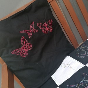 May include: A black pillow with pink geometric butterfly embroidery. The pillow is on a wooden bed frame with a white and black patchwork quilt.
