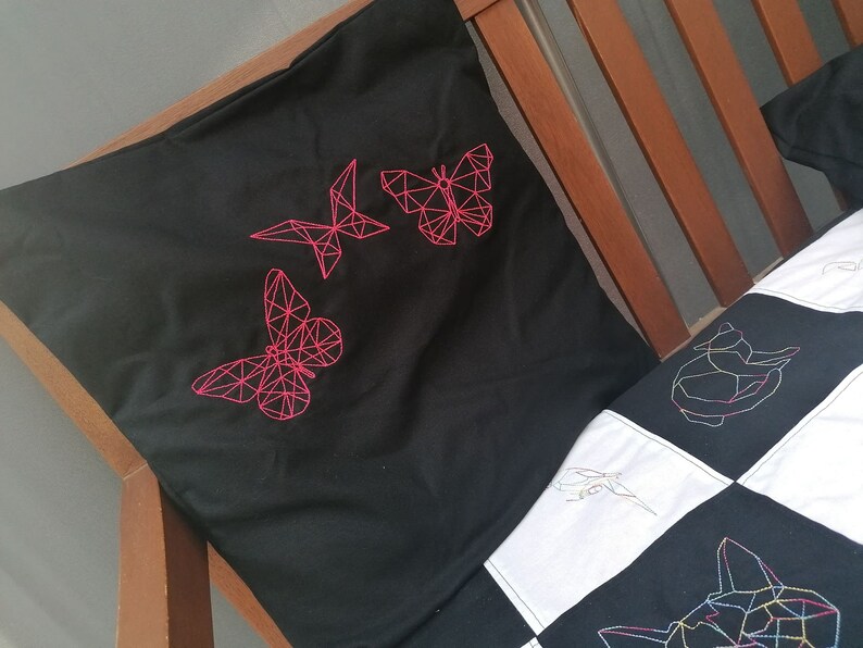 May include: A black pillow with pink geometric butterfly embroidery. The pillow is on a wooden bed frame with a white and black patchwork quilt.