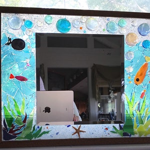 May include: A framed mirror with a mosaic design depicting an underwater scene. The frame is made of wood and the mosaic is made of glass tiles in shades of blue, green, and orange. The mirror is surrounded by a border of glass beads and there are two fish, one red and one orange, swimming in the scene.