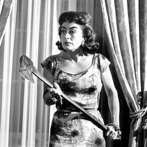 May include: Black and white image of a woman in a floral dress holding an axe. The dress has a pattern of large, stylized flowers. The woman has dark hair and is wearing jewelry. The image has a vintage feel.