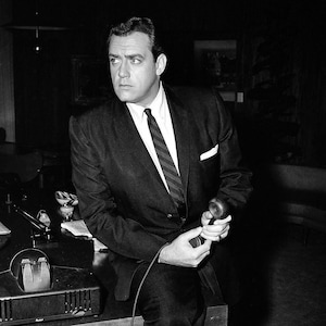 May include: A man in a suit and tie sits at a table, holding a microphone. He is looking off to the side, and his expression is serious. The table is covered in a dark cloth, and there is a microphone on the table in front of him.