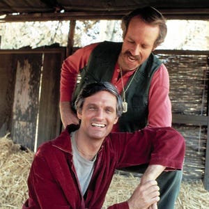 May include: A vintage photograph featuring two men in a rustic setting. One man is wearing a red velvet jumpsuit, while the other is in a red shirt, green vest, and necklace. The image has a watermark that reads "DO NOT COPY".