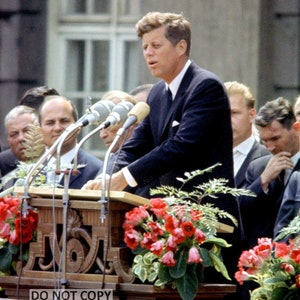 May include: A man in a dark suit speaks at a podium with microphones in front of him. He is surrounded by other men, and there are red flowers in front of the podium.